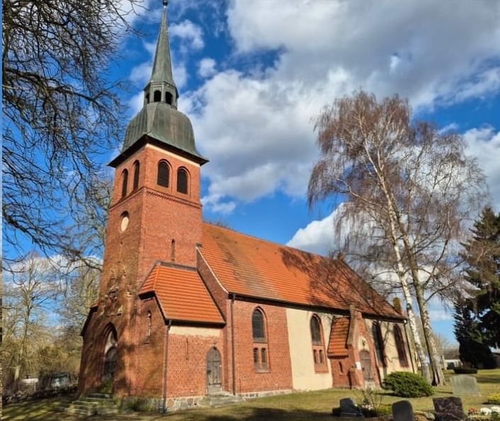 Die Backsteinkirche in Medow vor blauem Himmel mit Wolken, daneben eine Birke.