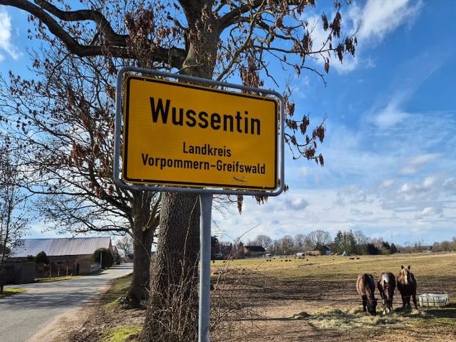 Ortseingangsschild Wussentin, dahinter eine Weide mit Pferden.